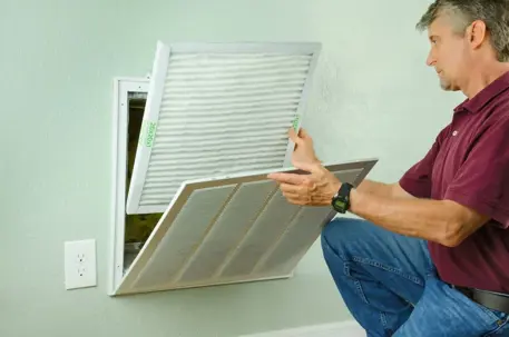 A man kneels, inserting a new, clean air filter into a wall-mounted return vent.