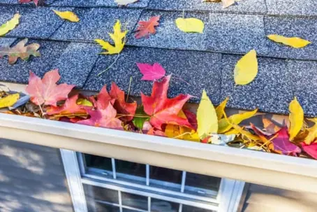 A home's gutter is overflowing with vibrant red, yellow, and orange fall leaves, illustrating common autumn home maintenance needs.
