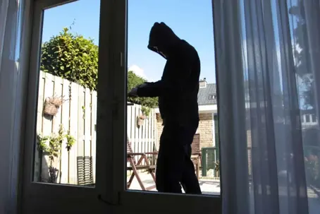 A dark silhouette of a person in a hood breaks into a house through a glass door. A sunny backyard with a fence is visible outside.