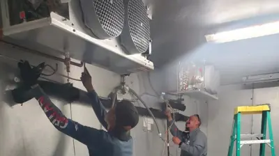 Two technicians work inside a walk-in cooler, connecting piping. Photo