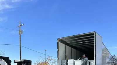 Two workers unload shiny metal ductwork from a semi-truck with a forklift on a bright, sunny day. Photo