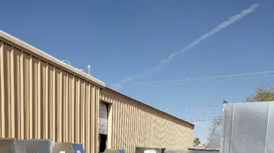 Stacks of galvanized metal ductwork are organized outside a beige industrial building under a blue sky, with a partial view of a Photo