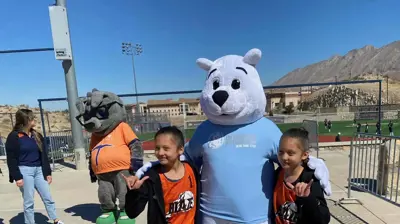 Two children in basketball jerseys pose with a white polar bear mascot and a grey rock mascot on an outdoor court. Photo