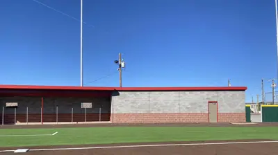 A sunny baseball field shows brown dirt in the foreground and green artificial turf. Photo