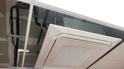 A white, square, ceiling-mounted air conditioning unit is partially installed in a dropped ceiling. Photo