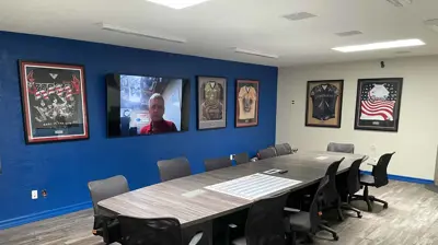 A modern conference room features a long table with ergonomic chairs, a blue accent wall, and a large TV displaying a man. Photo