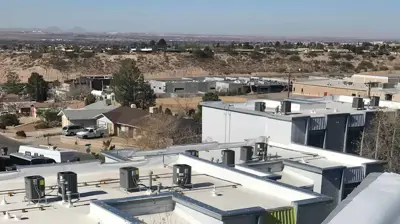 An elevated view of building rooftops with HVAC units, a tree, and a distant town under a clear blue sky. Photo