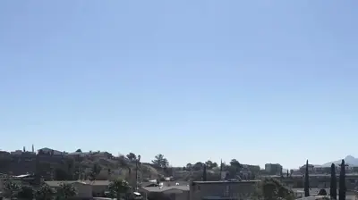 A series of modern buildings with green accents and flat roofs stretches towards distant hills under a clear blue sky. Photo
