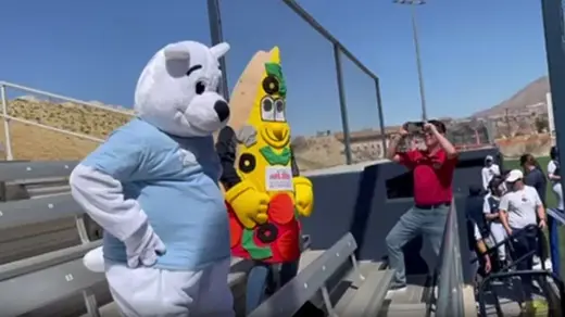 A white bear mascot and a colorful pizza mascot stand on bleachers as a man films them.